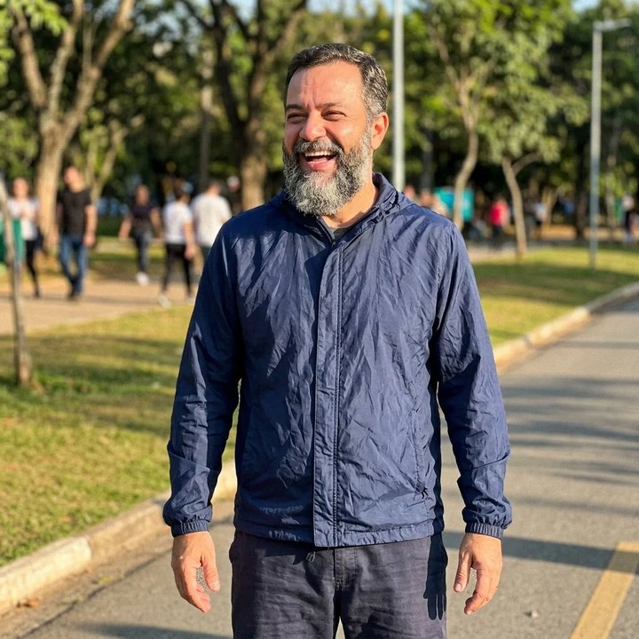 A laughing bearded man in a navy jacket standing on a sunny park path